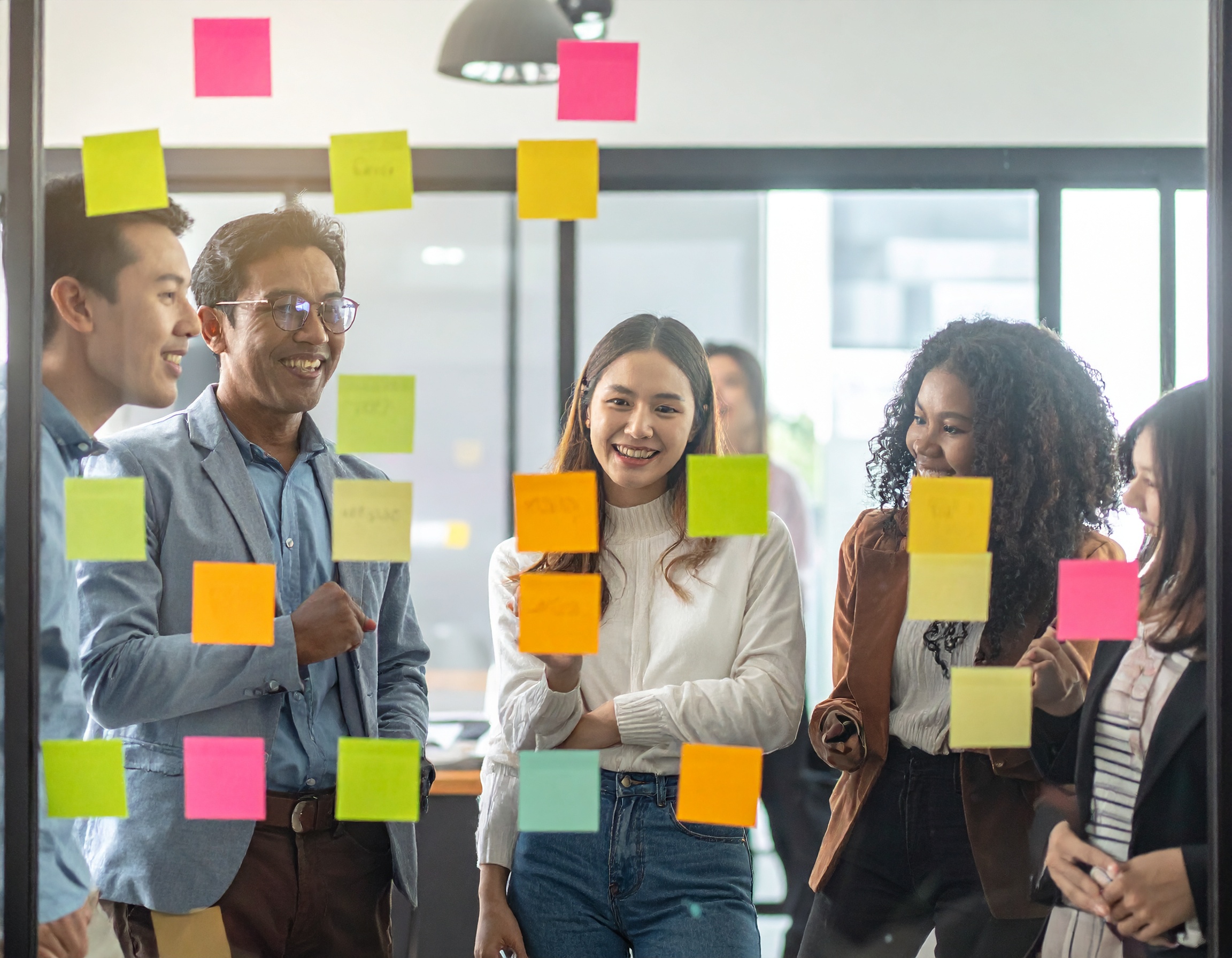 Grupo de jovens profissionais discutindo ideias em um escritório moderno, com post-its coloridos em uma parede de vidro, iluminação suave e ambiente colaborativo. Composição dinâmica destaca interação e diversidade, capturada num ângulo frontal com foco nítido nas expressões e nas cores vibrantes dos papéis adesivos.